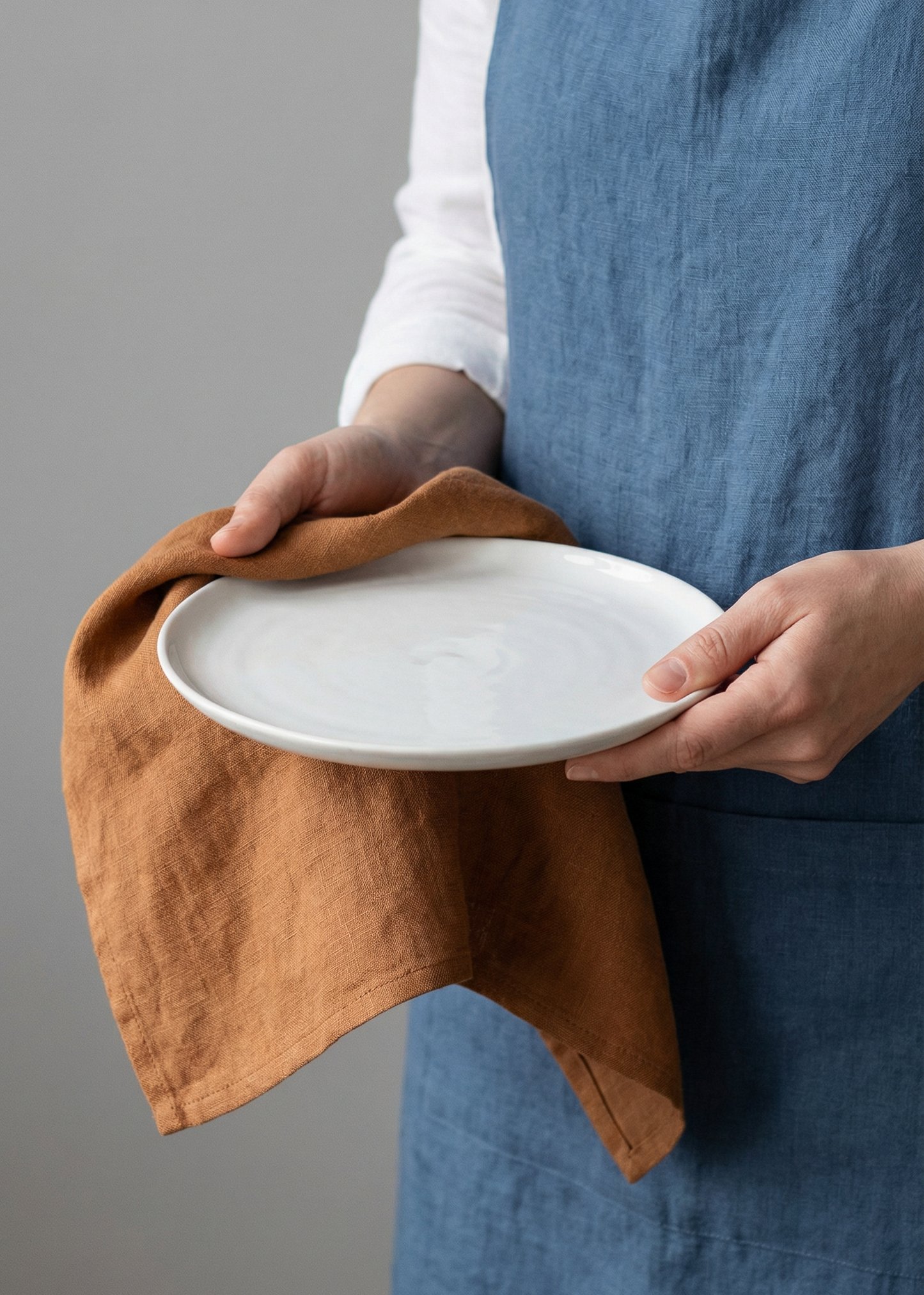 Person holding a white plate with a brown towel, wearing a blue apron against a gray background