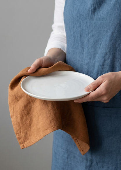 Person holding a white plate with a brown towel, wearing a blue apron against a gray background