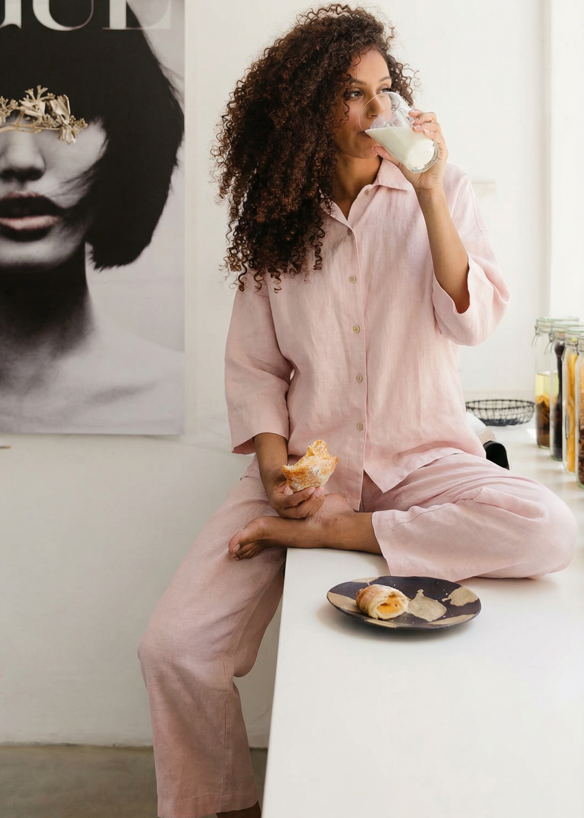 Woman in pink pajamas drinking milk and holding a donut in a cozy room.