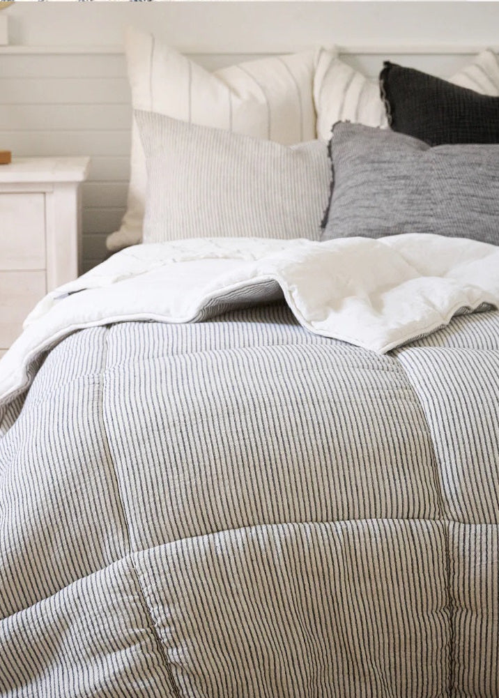 A bed with a striped reversible linen quilt in off white and ink color, paired with a pair of pillows, set in a bedroom interior.