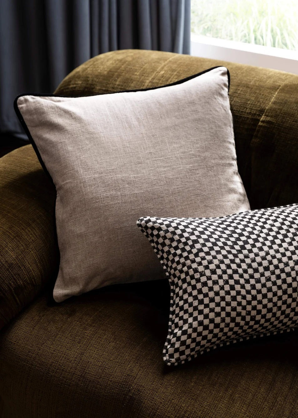 A linen cushion with black velvet piping on a sofa, with a checkered cushion beside it.
