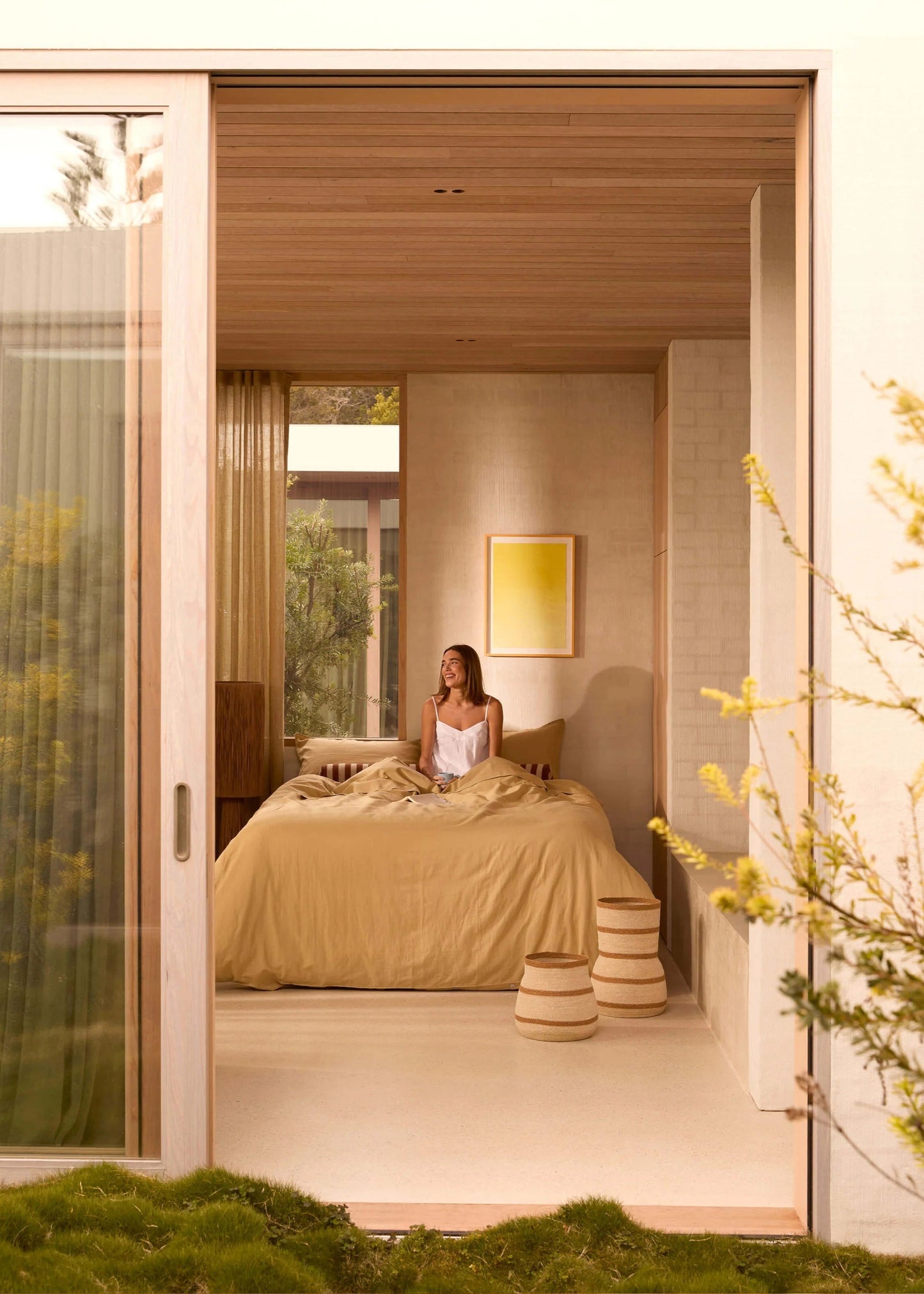 Woman sitting on a bed in a modern bedroom with a view of greenery outside.