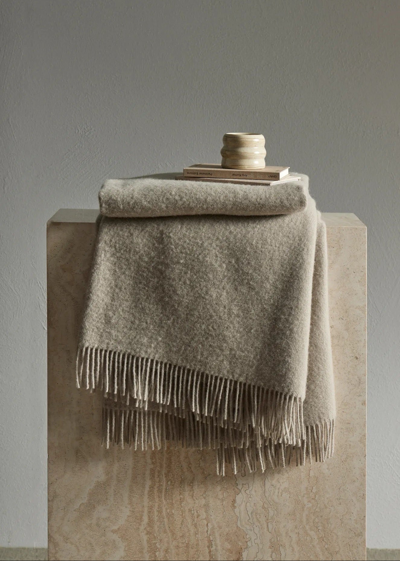 A folded beige throw blanket with fringe trim draped over a stone pedestal, with a small stack of books and a ceramic candle holder on top, against a neutral gray wall.