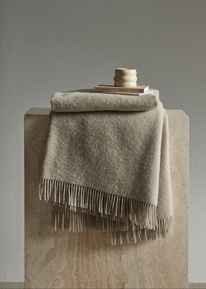 A folded beige throw blanket with fringe trim draped over a stone pedestal, with a small stack of books and a ceramic candle holder on top, against a neutral gray wall.