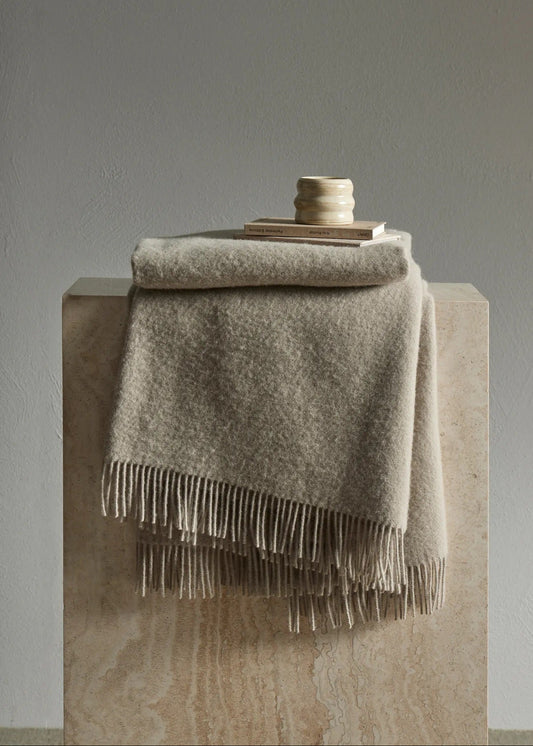 A folded beige throw blanket with fringe trim draped over a stone pedestal, with a small stack of books and a ceramic candle holder on top, against a neutral gray wall.