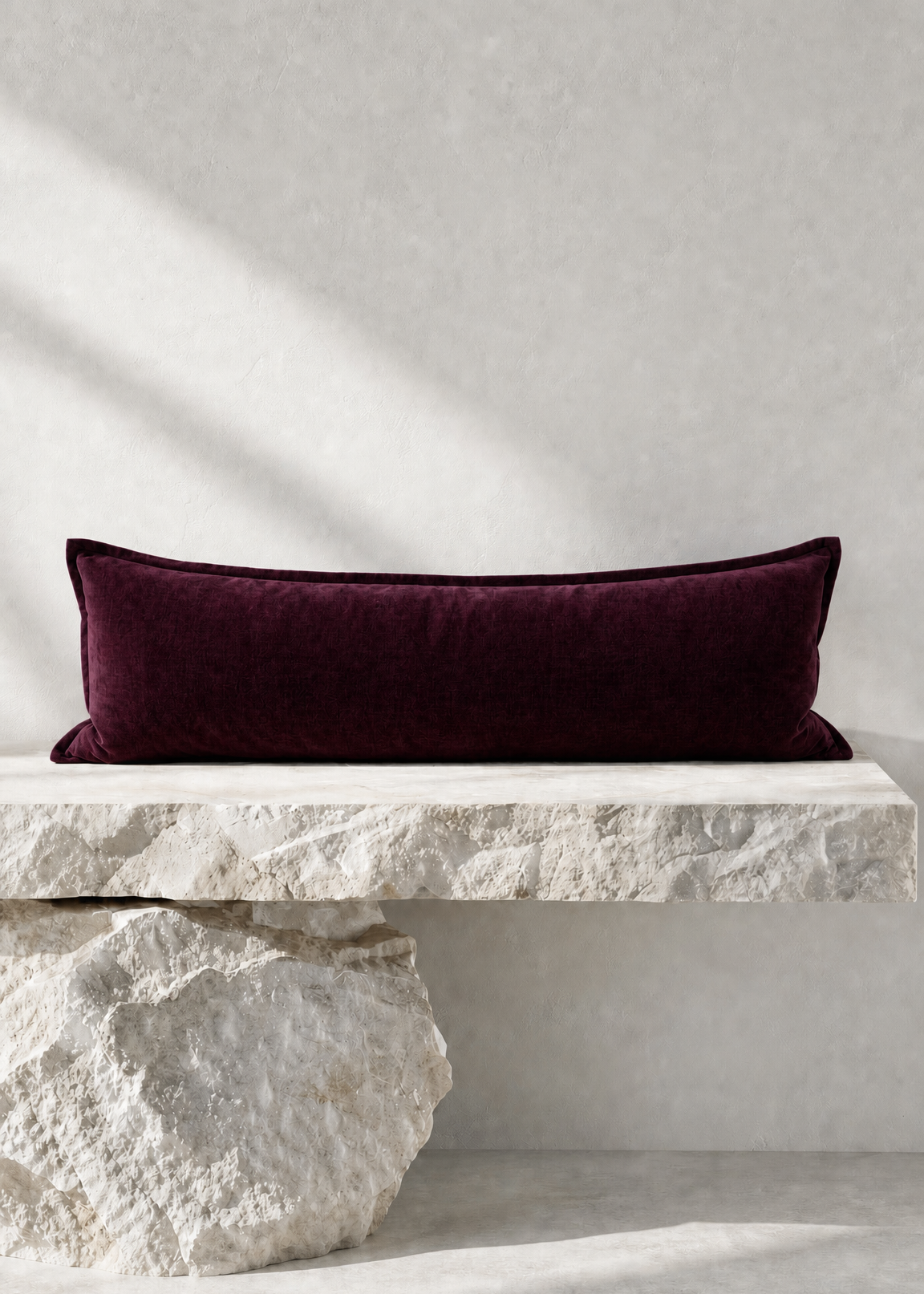 A long rectangular deep burgundy velvet pillow displayed on a white stone bench against a textured light gray wall with soft diagonal shadows.