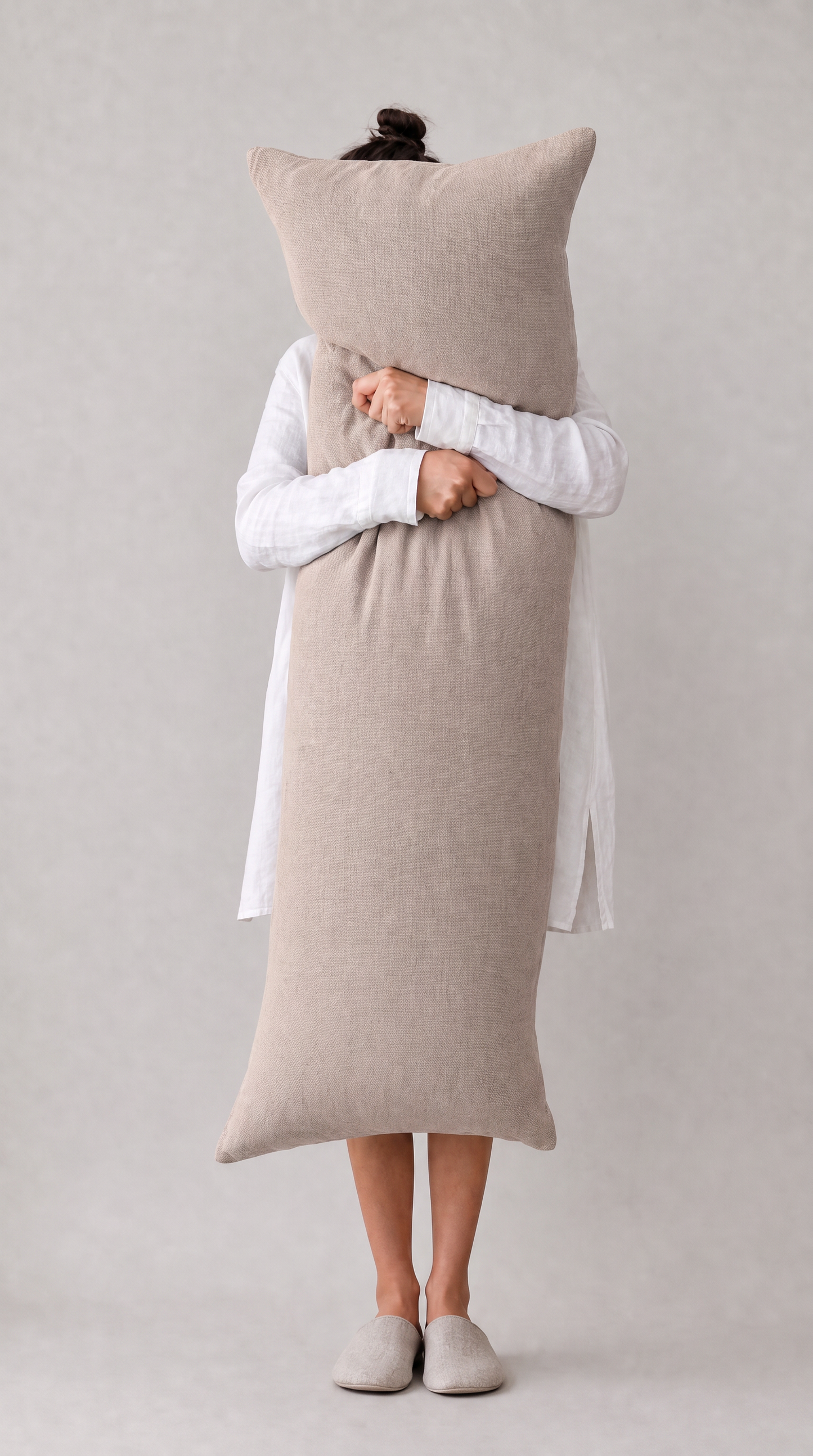 A person wearing a white long-sleeve dress and light-colored slippers holds a large, rectangular, ribbed beige pillow in front of their face against a neutral light gray background.