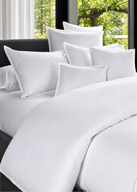 White bedding set with pillows on a bed in a room with large windows showing greenery outside.