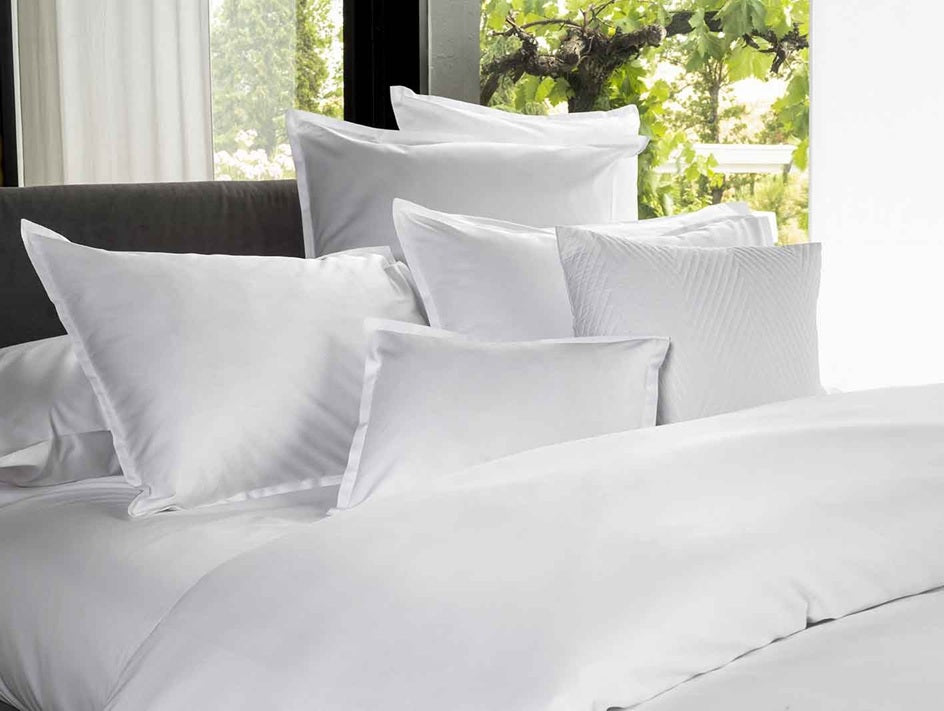 White bedding set with pillows on a bed in a room with a window view.