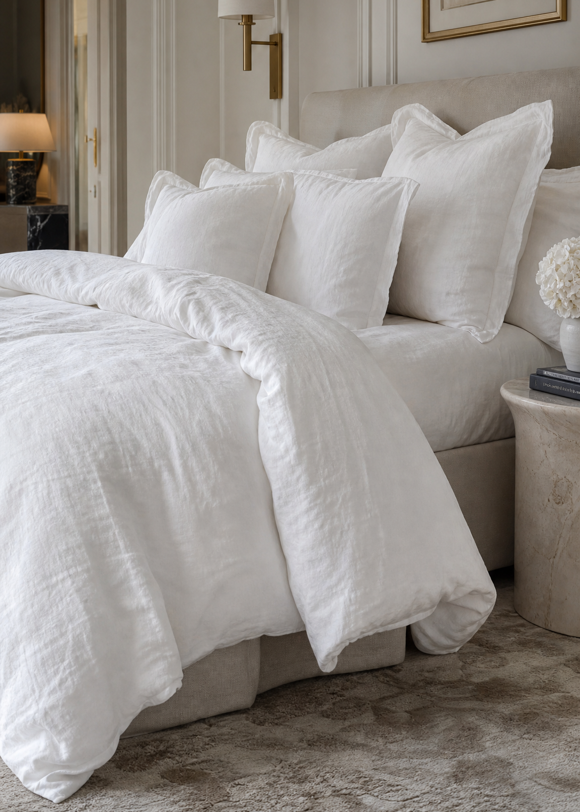 A neatly made bed in a softly lit bedroom, dressed in white textured bedding with multiple white pillows. The room has a beige upholstered headboard, a wall-mounted lamp, framed wall art, and a stone side table.