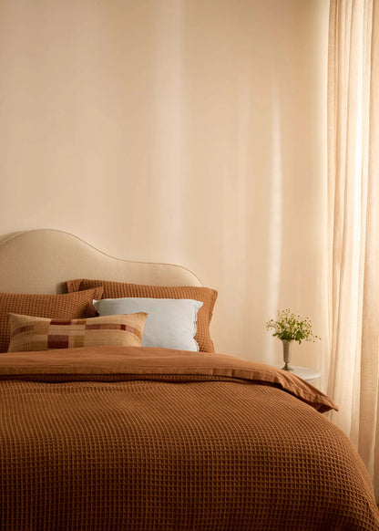 A cozy bedroom scene with a bed dressed in warm brown textured bedding, matching pillows, a cream upholstered headboard, sheer curtains, and a small vase of flowers on a bedside table.
