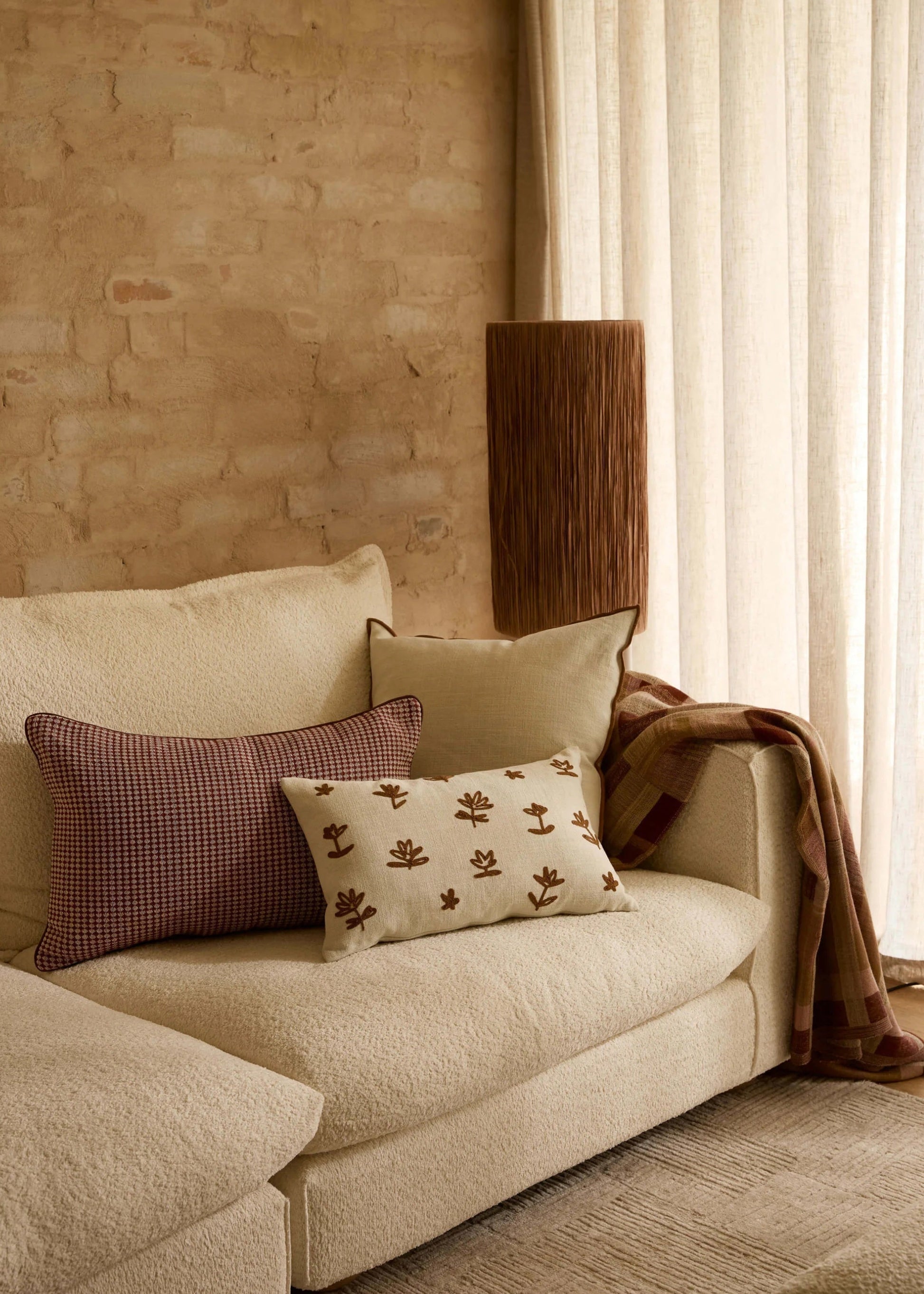 A cozy living room corner featuring a cream bouclé sofa with decorative throw pillows, a draped brown throw blanket, a tall textured wooden floor lamp, and a warm brick wall next to sheer curtains.
