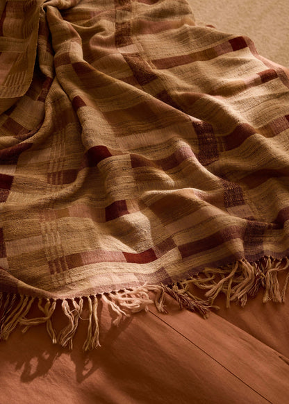 A close-up of a woven throw blanket with fringed edges, draped over a wooden surface. The blanket has a plaid pattern in warm beige, brown, and muted red tones.