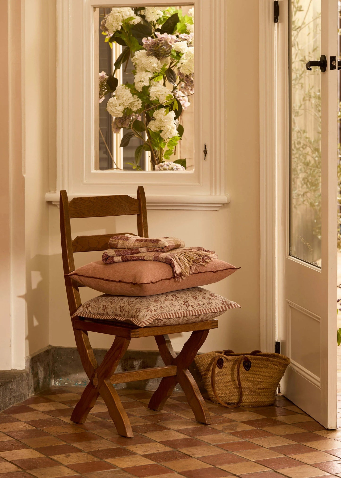 A rustic wooden folding chair in a warm, sunlit entryway with terracotta tile flooring, holding stacked cushions, a folded throw blanket, and a woven basket bag.
