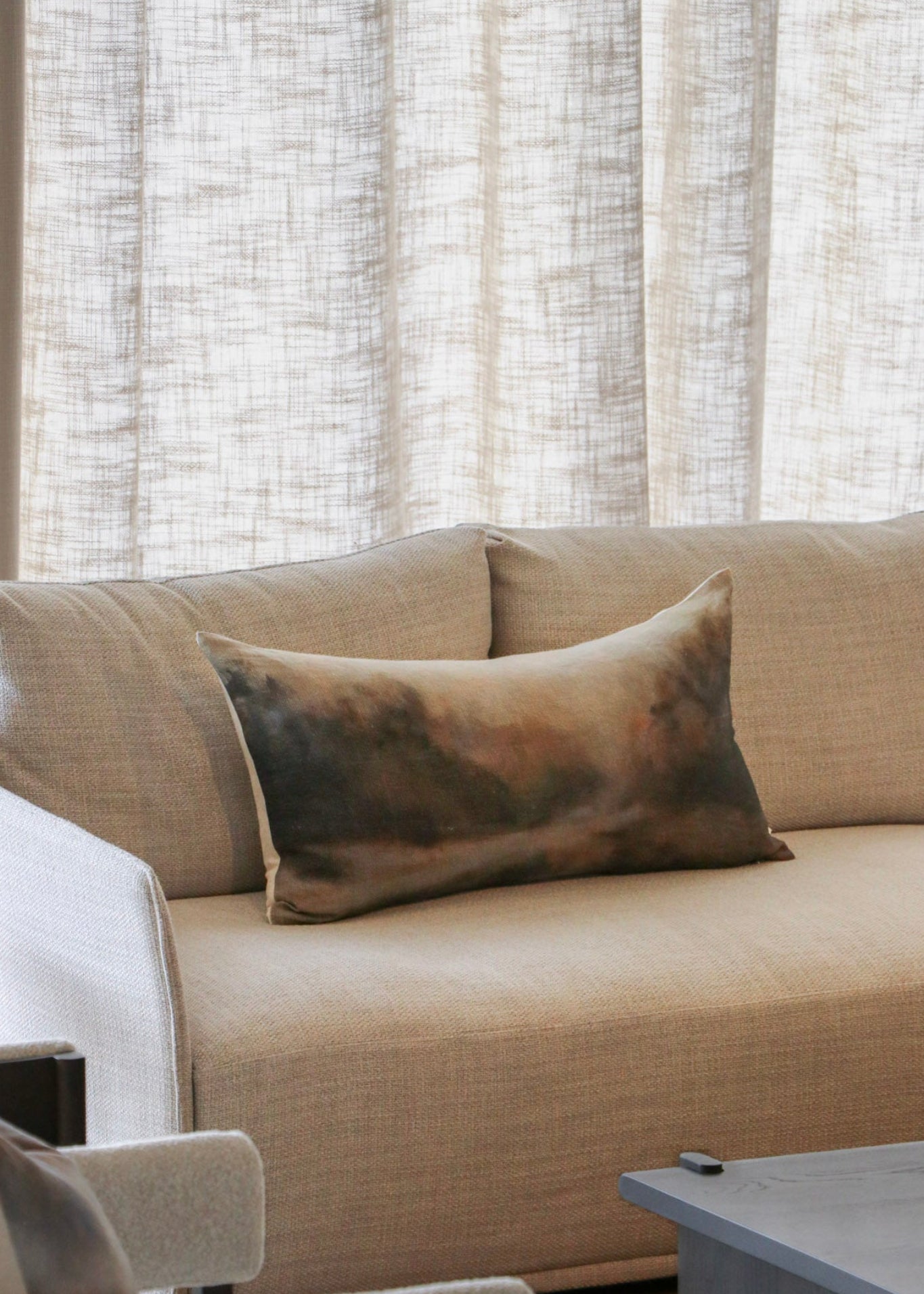A beige upholstered sofa in a living room setting, styled with a rectangular throw pillow featuring a mottled brown, gold, and beige pattern. The background consists of textured light beige curtains.