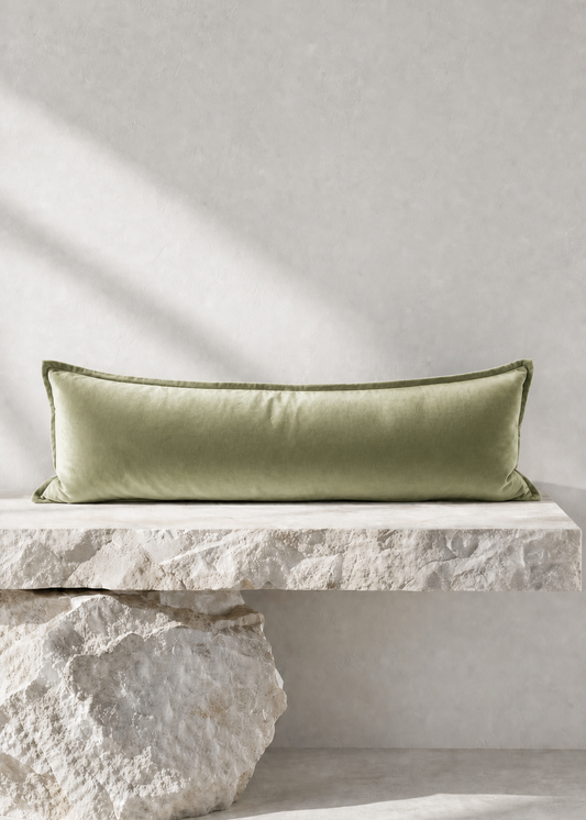 A long rectangular sage green velvet lumbar pillow displayed on a rustic stone bench against a textured light gray wall.