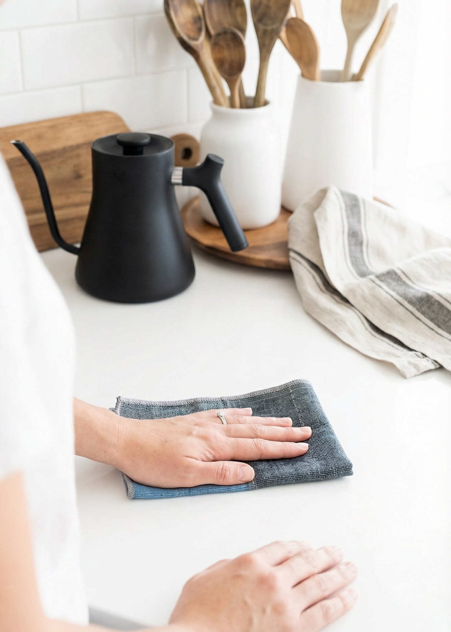 Person cleaning a kitchen counter with a black coffee pot and wooden utensils in the background.