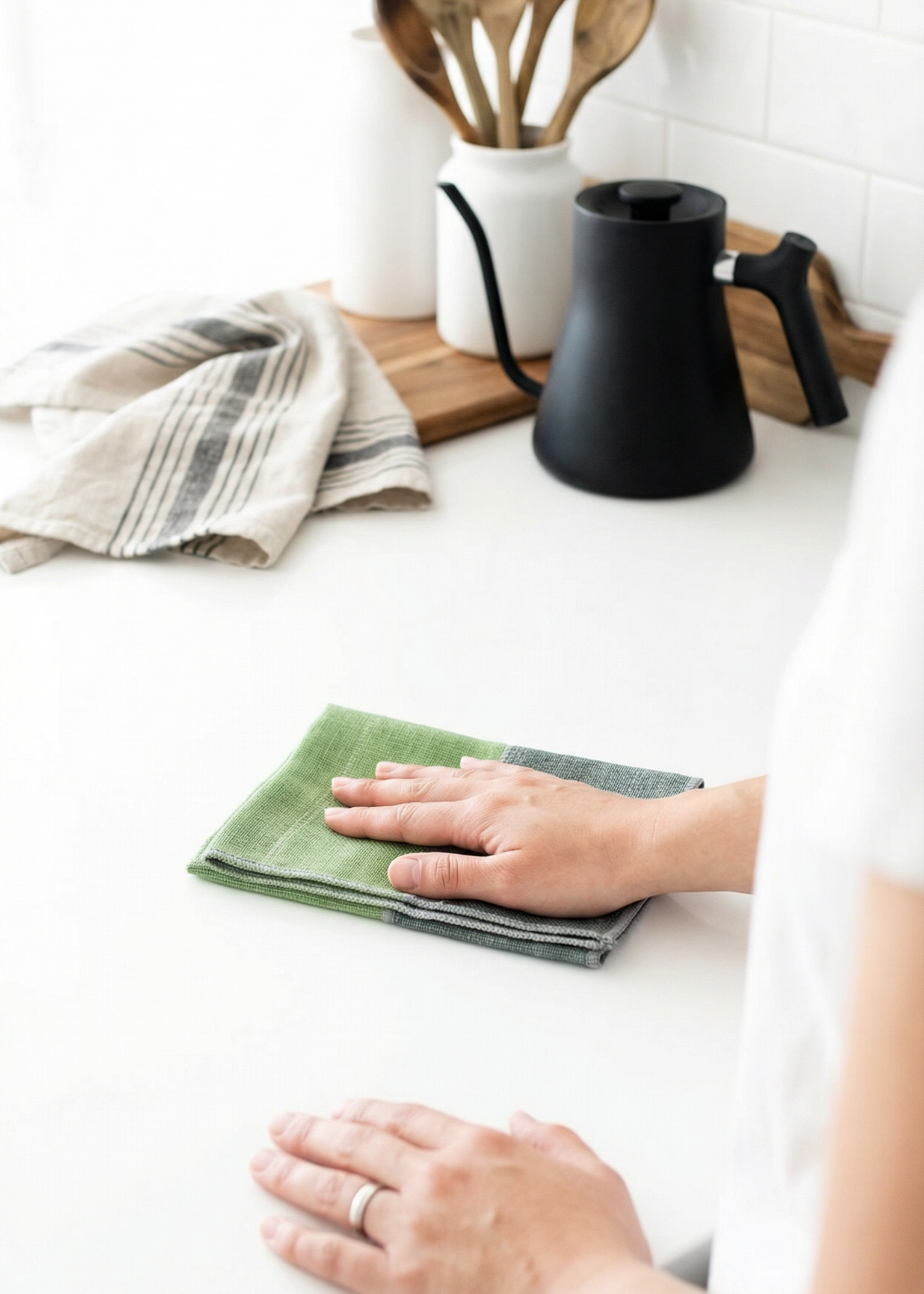 Person cleaning a kitchen counter with a green cloth, surrounded by kitchen utensils and a coffee maker.