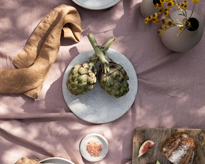 Artichokes on a white plate with bread, figs, and a vase of flowers on a pink surface
