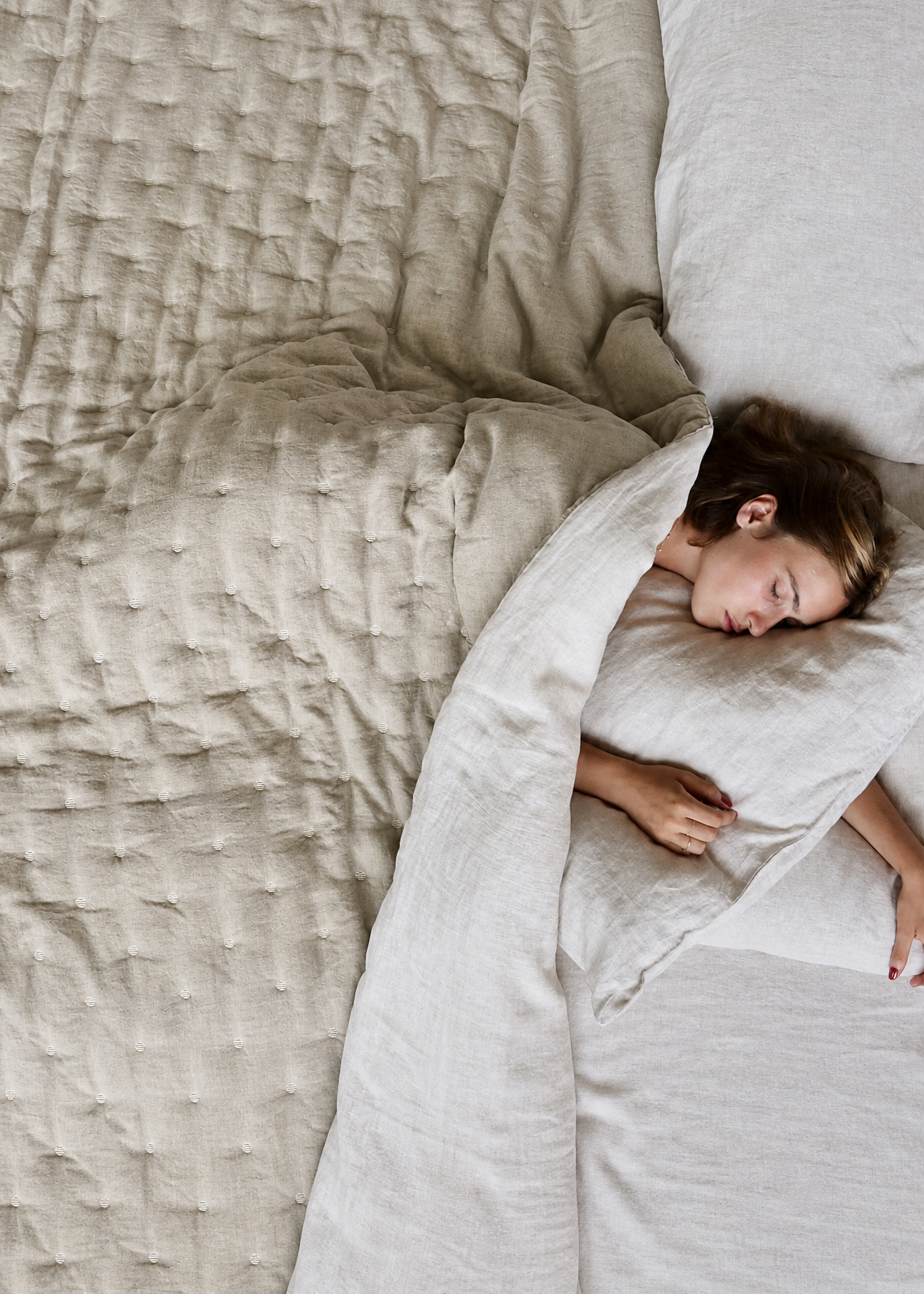 Person sleeping under a light-colored comforter on a bed with neutral bedding.