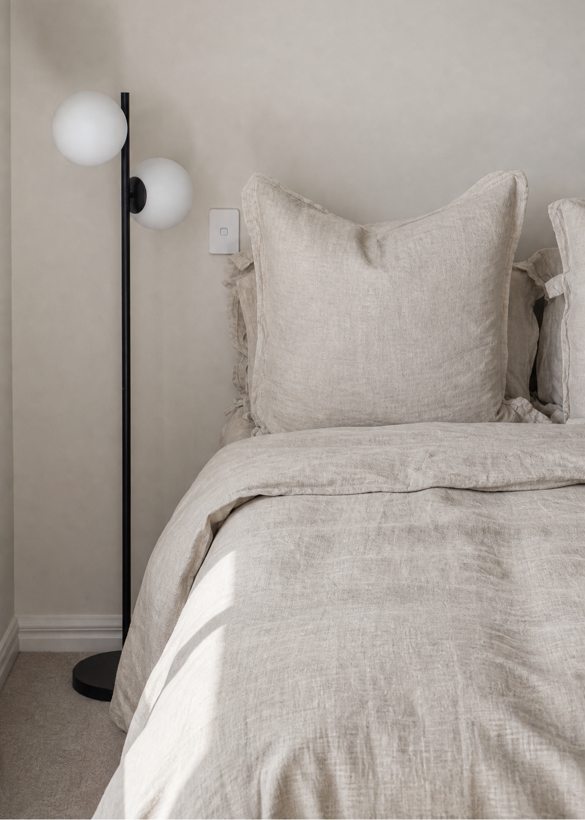 A cozy bedroom scene featuring a neatly made bed with textured beige linen bedding and matching pillows, next to a black floor lamp with two white globe shades against a neutral beige wall.