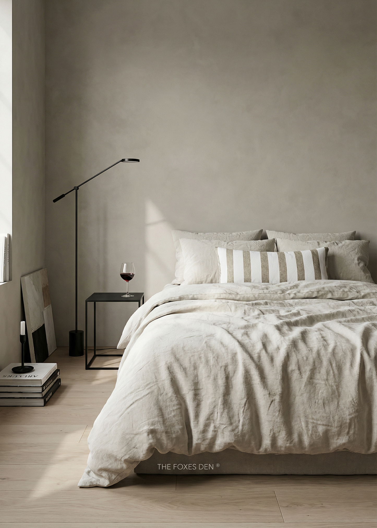 A styled bedroom scene showing a bed dressed in light beige and white bedding, with a striped accent pillow, a black floor lamp, a small side table with a wine glass, and a textured plaster wall in the background. The text 'THE FOXES DEN' is visible at the bottom of the bed.