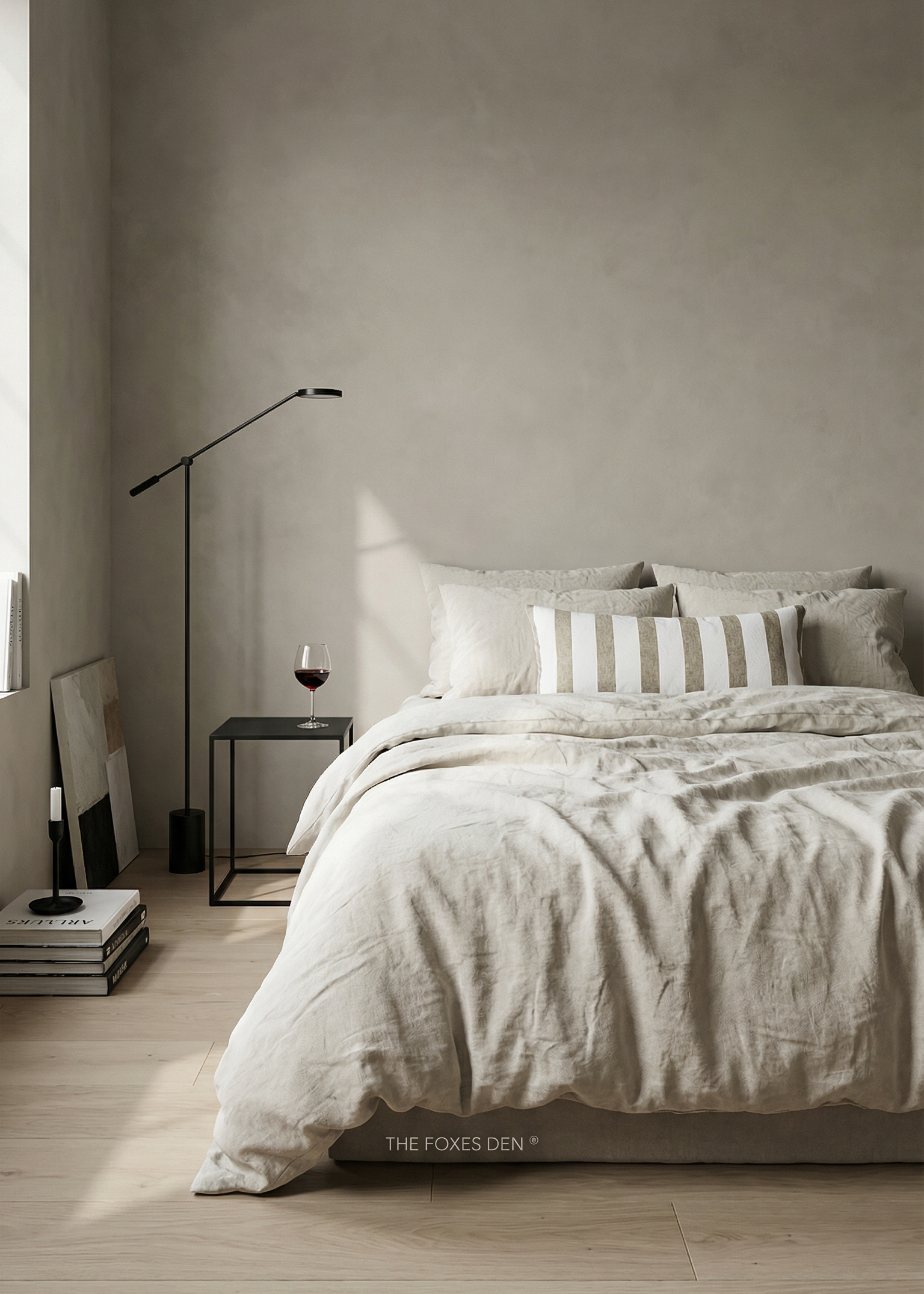 A styled bedroom scene showing a bed dressed in light beige and white bedding, with a striped accent pillow, a black floor lamp, a small side table with a wine glass, and a textured plaster wall in the background. The text 'THE FOXES DEN' is visible at the bottom of the bed.
