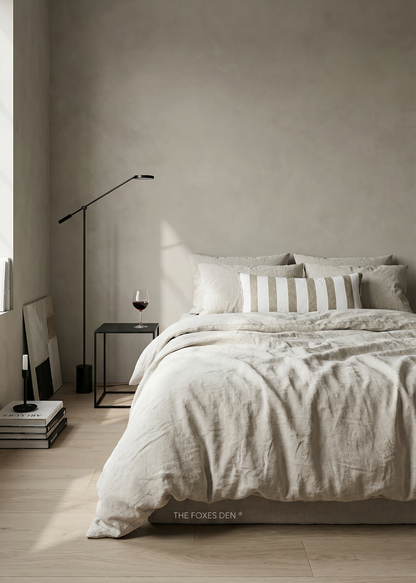 A styled bedroom scene showing a bed dressed in light beige and white bedding, with a striped accent pillow, a black floor lamp, a small side table with a wine glass, and a textured plaster wall in the background. The text 'THE FOXES DEN' is visible at the bottom of the bed.