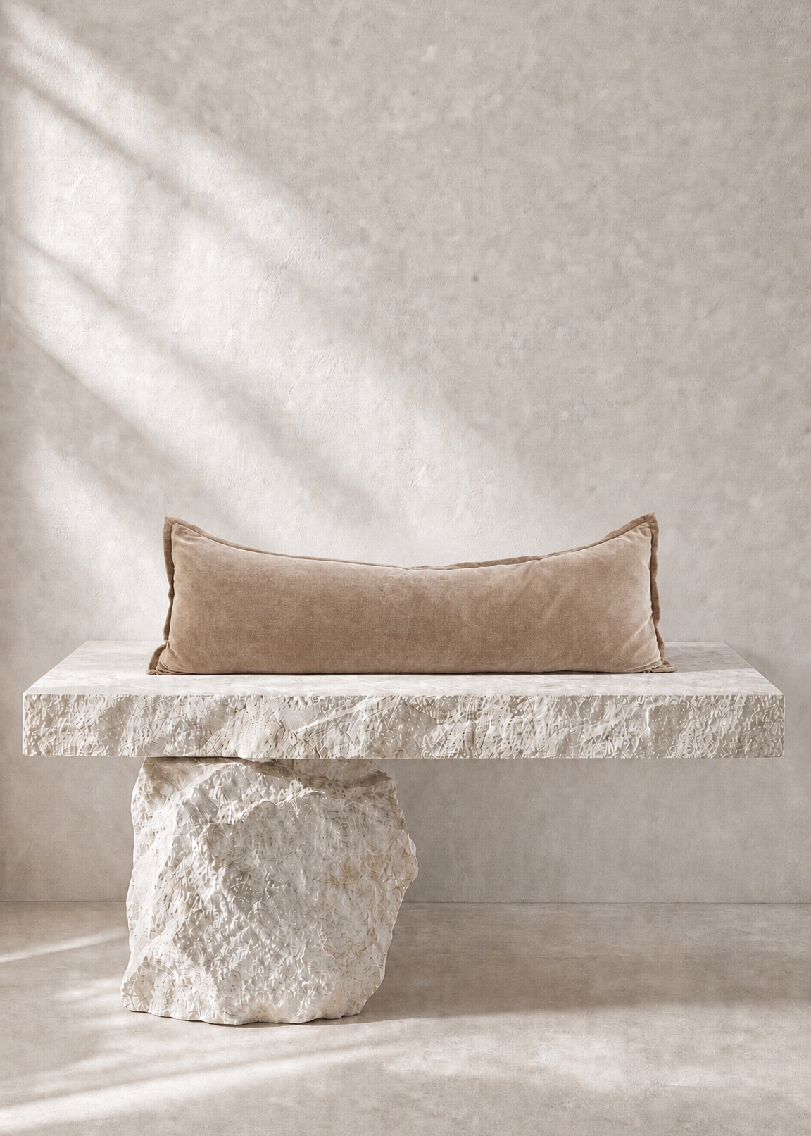 A rectangular beige pillow with a textured surface, resting on a rustic stone bench against a textured beige wall with soft diagonal shadows.