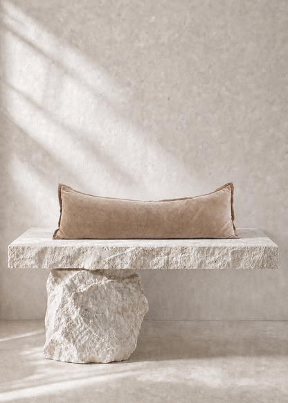 A rectangular beige pillow with a textured surface, resting on a rustic stone bench against a textured beige wall with soft diagonal shadows.