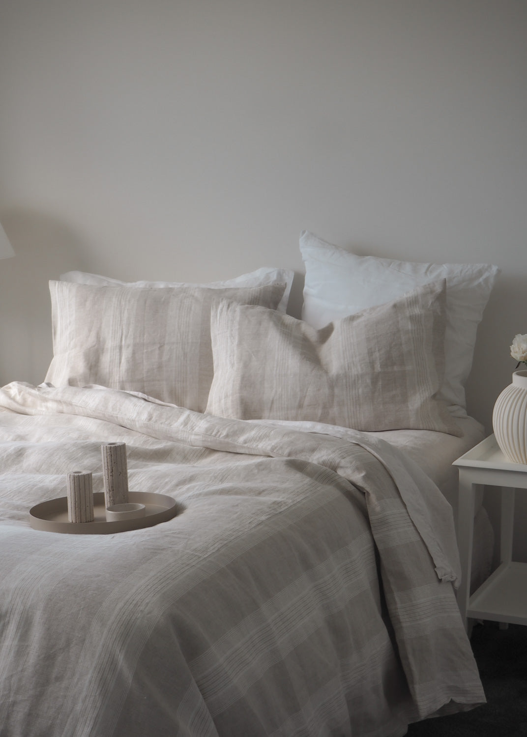 Neatly made bed with light-colored bedding and pillows against a plain wall.