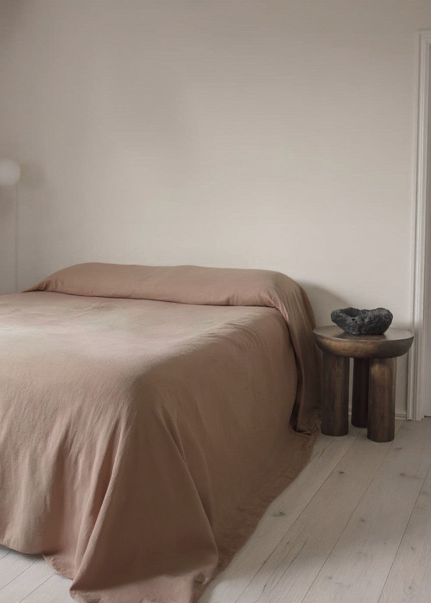 Bed with brown bedding in a minimalistic room with a wooden stool and decorative bowl.
