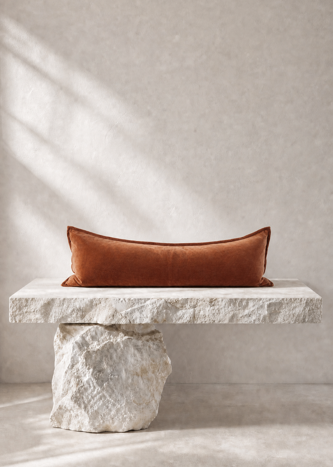 A rectangular rust-orange velvet pillow displayed on a rustic white stone bench against a textured beige wall with soft diagonal shadows.