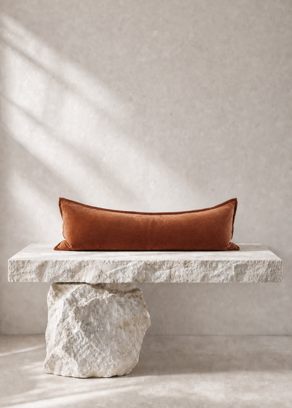 A rectangular rust-orange velvet pillow displayed on a rustic white stone bench against a textured beige wall with soft diagonal shadows.