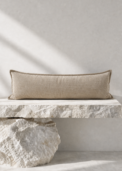 A rectangular beige linen lumbar pillow with a subtle stitched border, resting on a rustic white stone bench against a textured light gray wall with soft diagonal shadows.
