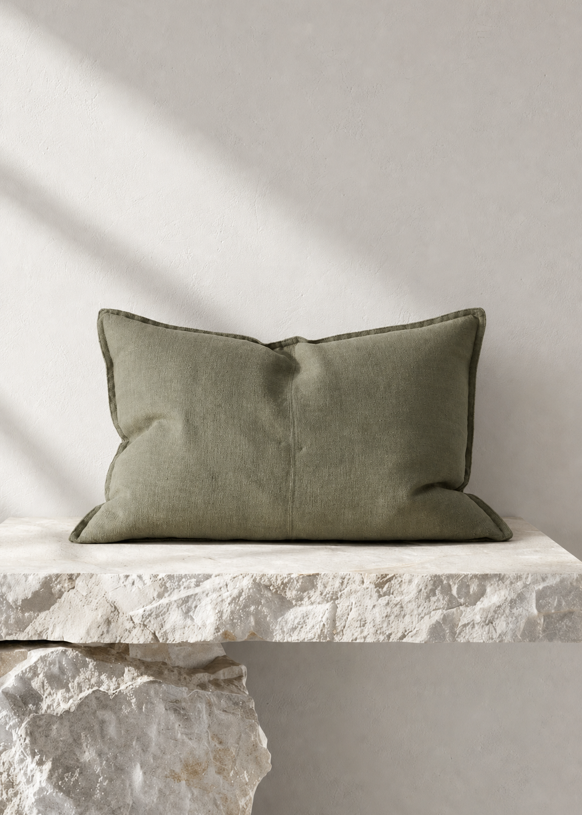 An olive green rectangular throw pillow with piped edges, resting on a rustic white stone ledge against a textured light gray wall with soft diagonal shadows.