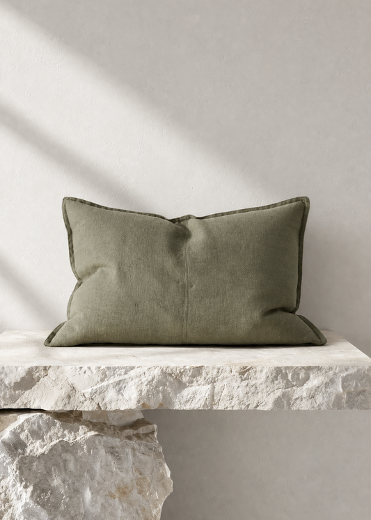 An olive green rectangular throw pillow with piped edges, resting on a rustic white stone ledge against a textured light gray wall with soft diagonal shadows.