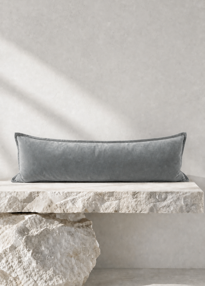 A long rectangular gray velvet lumbar pillow resting on a rustic stone bench against a textured light gray wall.
