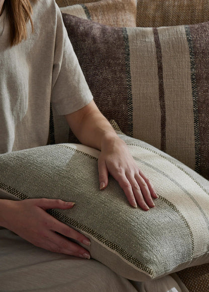 A person in a beige linen shirt holds a textured cream pillow with woven stripes, with another striped pillow and a sofa in the background.