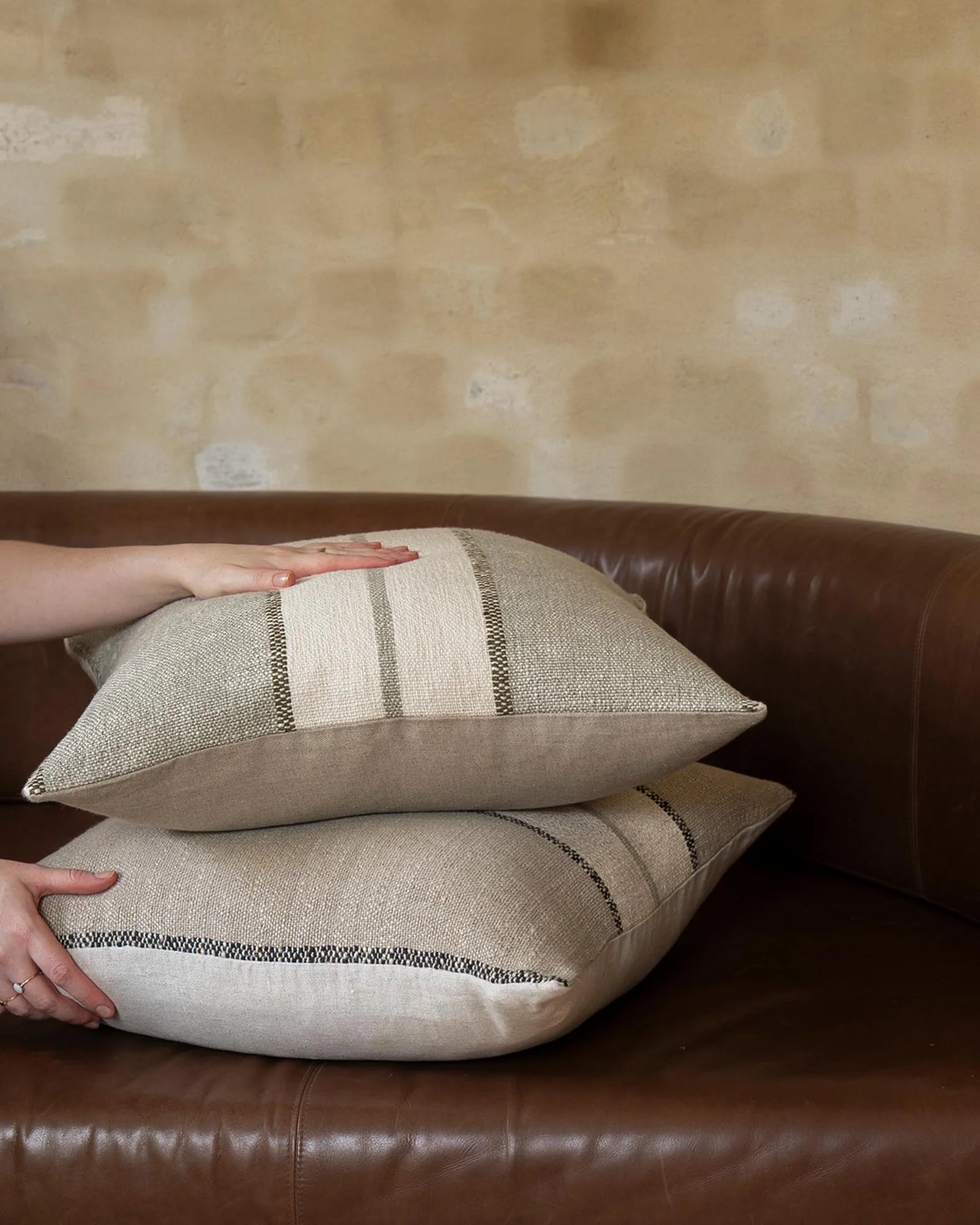 Two beige woven throw pillows with striped detailing are stacked on a brown leather sofa, with hands adjusting them against a textured beige wall.