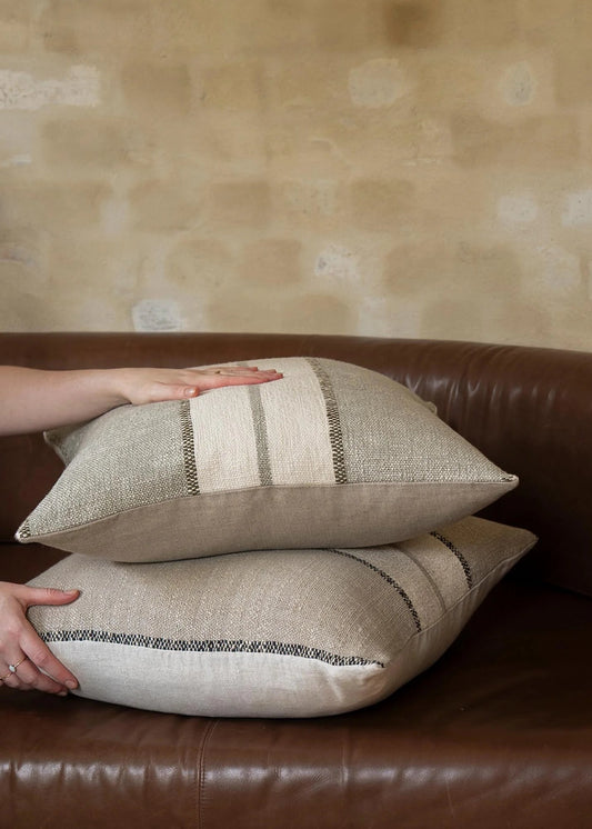Two beige woven throw pillows with striped detailing are stacked on a brown leather sofa, with hands adjusting them against a textured beige wall.