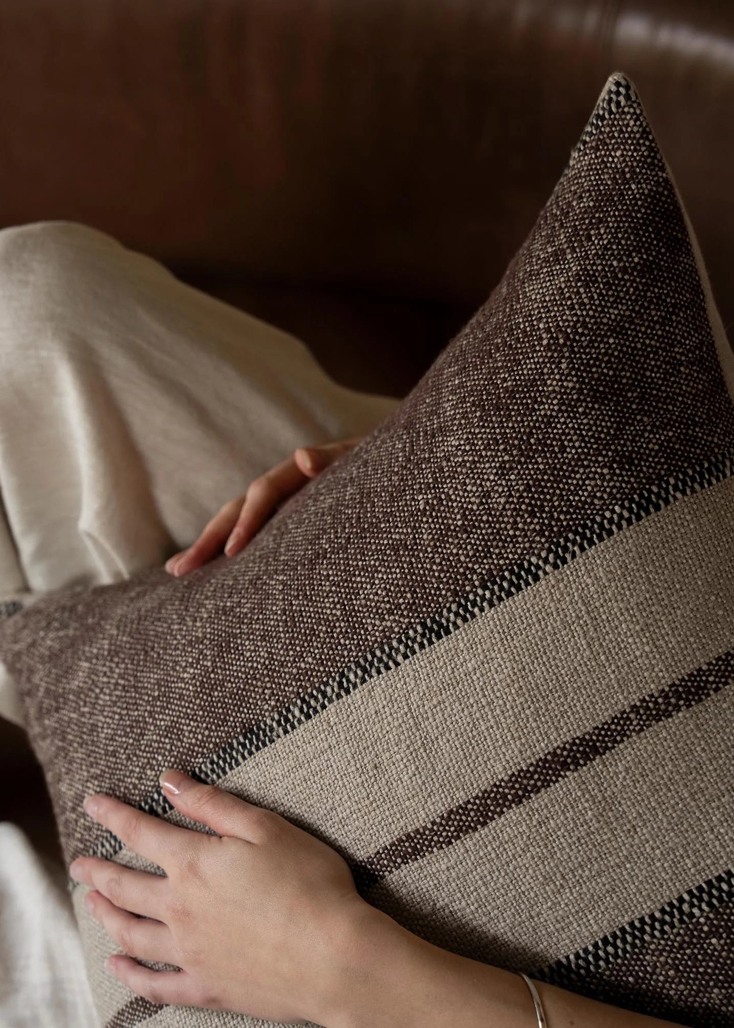 A close-up of a textured woven throw pillow in brown and beige tones, being held by a person sitting on a brown leather sofa.