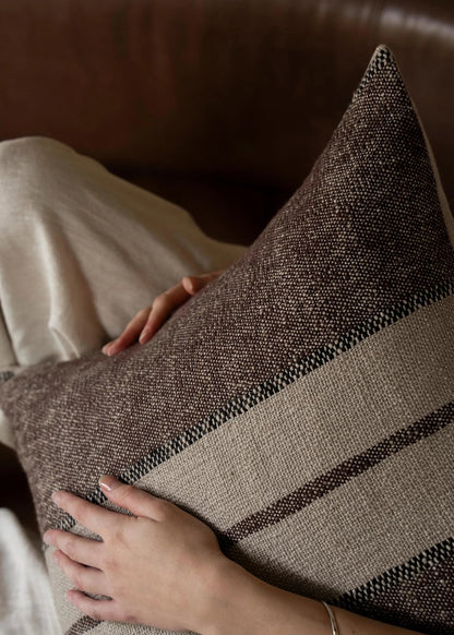 A close-up of a textured woven throw pillow in brown and beige tones, being held by a person sitting on a brown leather sofa.
