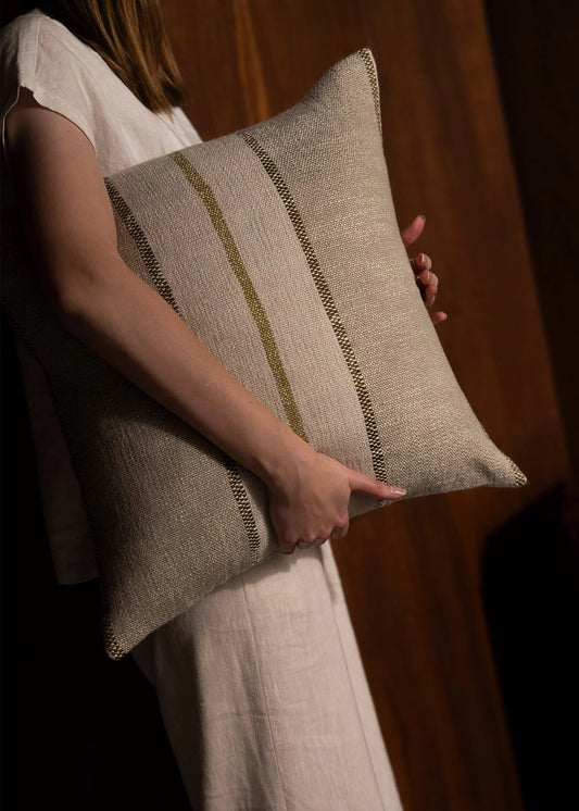 A person in a white dress holds a square woven throw pillow with a textured beige linen-like fabric and two vertical olive-green stripes.