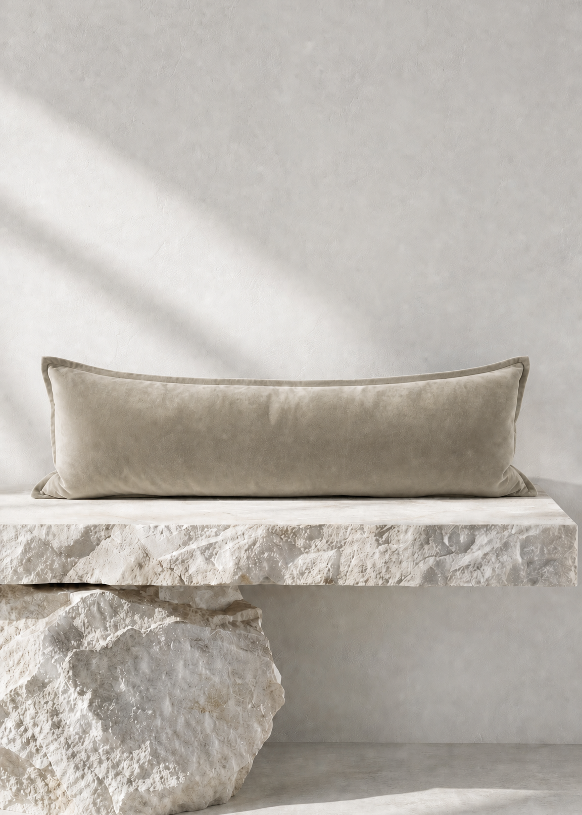 A long rectangular lumbar pillow in a textured taupe-beige velvet fabric, resting on a floating white stone bench against a neutral concrete wall.