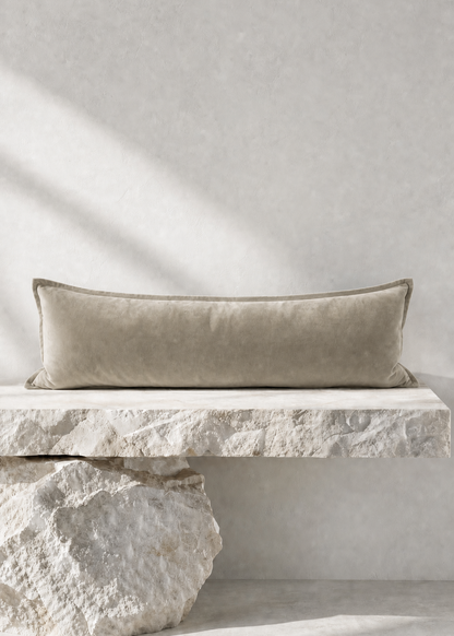 A long rectangular lumbar pillow in a textured taupe-beige velvet fabric, resting on a floating white stone bench against a neutral concrete wall.