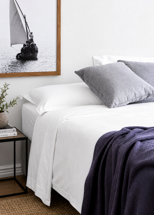 A styled bedroom scene showing a neatly made bed with white bedding, two gray pillows, a dark navy throw blanket, a small black side table with books and a plant, and a framed black-and-white sailboat print on a white wall.