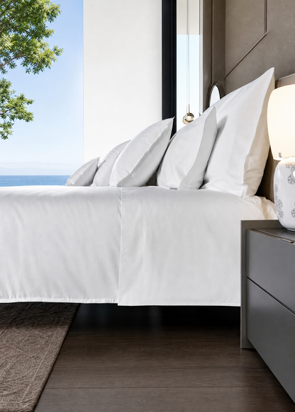 A luxury bedroom scene featuring a bed dressed in crisp white linens and multiple pillows, positioned next to a dark wood nightstand with a lit lamp. The room has dark wood flooring, a textured rug, and a large window looking out to the blue ocean and sky.