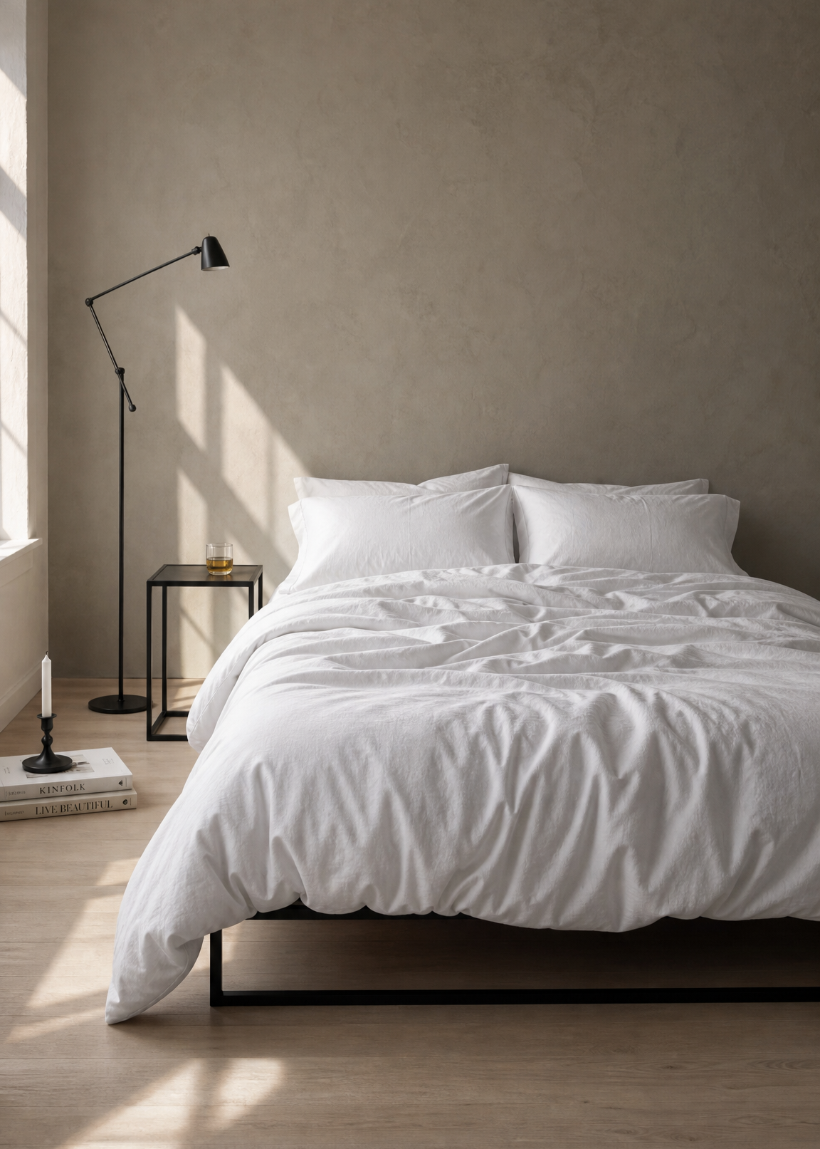 A minimalist bedroom scene featuring a bed dressed in crisp white bedding, set against a textured warm gray wall with soft sunlight streaming across the floor and wall.