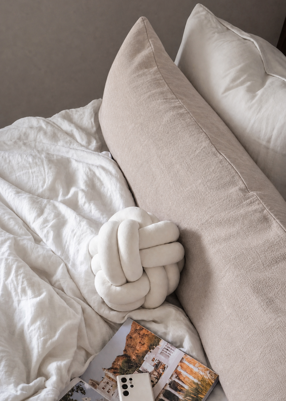 A styled bedroom scene with white bedding, a large beige pillow, a knotted white throw pillow, and a magazine with a white smartphone resting on top.